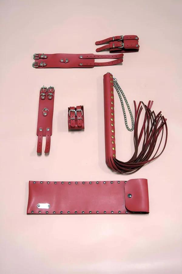 Red vegan leather accessory set with wide studded belt, double-buckle wrist cuffs with silver D-rings, and multi-tassel flogg