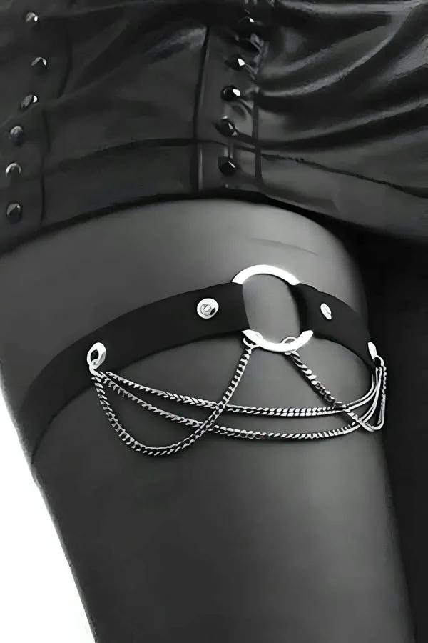 Black faux leather garter harness with silver O-ring and triple chain draping worn on upper thigh over sheer black stockings