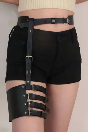 Black faux leather garter harness with adjustable straps and silver buckles worn on a person's thigh.