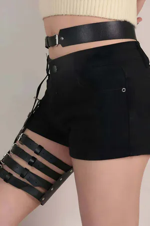 Black faux leather garter harness with adjustable straps and silver buckles worn on a person's thigh.
